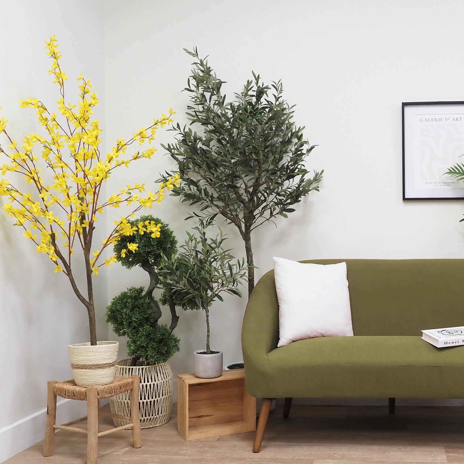 Forsythia artificiel jaune 150cm dans un cache-pot posé sur un tabouret à côté d'autres arbres artificiels et un canapé